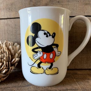 K&ouml;nnte beinhalten: Wei&szlig;e Keramiktasse mit einem Mickey Mouse-Design. Die Cartoonfigur ist in Schwarz, Rot, Gelb und Wei&szlig; gehalten und vor einem gelben Kreis platziert. Die Tasse hat einen gebogenen Griff und eine gl&auml;nzende Oberfl&auml;che.