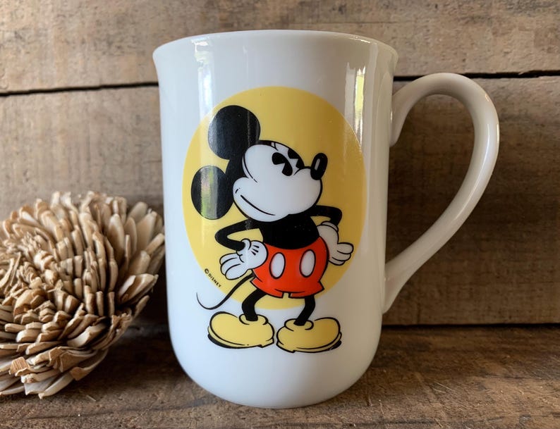 K&ouml;nnte beinhalten: Wei&szlig;e Keramiktasse mit einem Mickey Mouse-Design. Die Cartoonfigur ist in Schwarz, Rot, Gelb und Wei&szlig; gehalten und vor einem gelben Kreis platziert. Die Tasse hat einen gebogenen Griff und eine gl&auml;nzende Oberfl&auml;che.