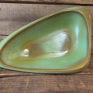May include: A mid-century modern, asymmetrical ceramic dish in shades of green and gold. The dish has a smooth, glossy finish and a shallow, curved interior. The exterior has a slightly textured appearance. The dish is displayed on a rustic wooden surface.