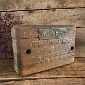 May include: A rectangular wooden block featuring a weathered surface with two circular holes. A metal plate is attached to the front, embossed with the text A.75-. The wood also displays the stamped alphanumeric sequence C123696.