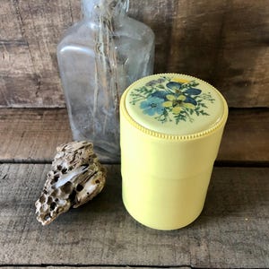 Peut inclure: Un récipient jaune vintage avec un couvercle floral orné de fleurs bleues et jaunes. Une bouteille en verre transparent et un morceau de bois flotté sont en arrière-plan. Le récipient est cylindrique et a un bord perlé.