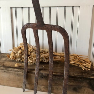 May include: A rustic, antique garden fork with four tines and a long handle. The metal tool has a weathered, brown finish, suggesting age and use. The fork is displayed against a white-painted wood background.