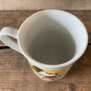 K&ouml;nnte beinhalten: Wei&szlig;er Keramikbecher mit gebogenem Henkel. Der Becher zeigt ein Cartoon-Charakter-Design mit schwarzen, roten und gelben Akzenten. Der Becher steht auf einer rustikalen Holzoberfl&auml;che.