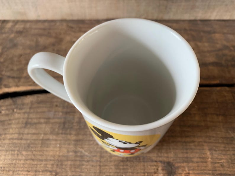 K&ouml;nnte beinhalten: Wei&szlig;er Keramikbecher mit gebogenem Henkel. Der Becher zeigt ein Cartoon-Charakter-Design mit schwarzen, roten und gelben Akzenten. Der Becher steht auf einer rustikalen Holzoberfl&auml;che.
