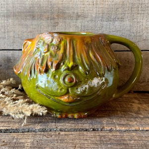 May include: A ceramic mug with a fish-like design. The mug is primarily green with orange and yellow accents. It has a handle and a detailed face with a smiling expression. The mug sits on a wooden surface.