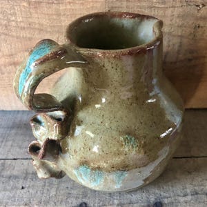 May include: A handcrafted ceramic jug with a distinctive handle. The jug has a speckled, earthy green glaze with turquoise and brown highlights. The handle has a unique, sculptural form, enhancing its artistic quality. Ideal for serving drinks, this jug is a functional art piece.