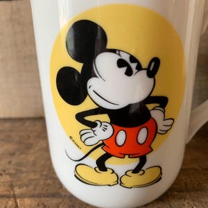 K&ouml;nnte beinhalten: Wei&szlig;e Keramiktasse mit Mickey Mouse-Design. Die Cartoonfigur ist in Schwarz, Rot, Gelb und Wei&szlig; gehalten und befindet sich auf einem gelben Kreis. Die Tasse hat einen Henkel und ist f&uuml;r Getr&auml;nke geeignet.