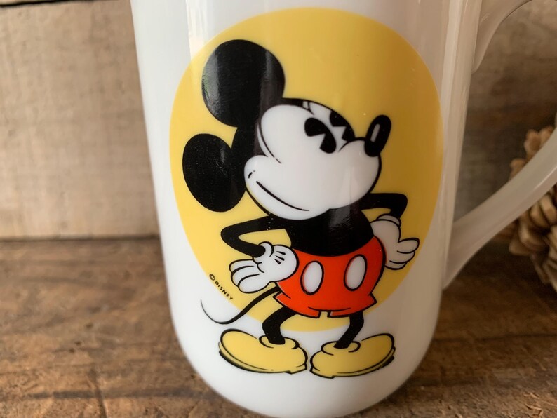 K&ouml;nnte beinhalten: Wei&szlig;e Keramiktasse mit Mickey Mouse-Design. Die Cartoonfigur ist in Schwarz, Rot, Gelb und Wei&szlig; gehalten und befindet sich auf einem gelben Kreis. Die Tasse hat einen Henkel und ist f&uuml;r Getr&auml;nke geeignet.