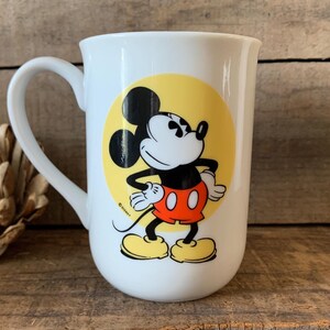 K&ouml;nnte beinhalten: Wei&szlig;e Keramiktasse mit Mickey Mouse-Design. Die Tasse hat einen gro&szlig;en Henkel und einen gelben Kreis mit Mickey Mouse in seiner ikonischen Pose, mit roter Hose und gelben Schuhen. Ein Disney-Lizenzprodukt.