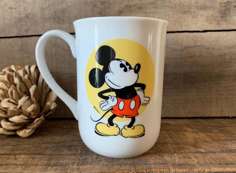 K&ouml;nnte beinhalten: Wei&szlig;e Keramiktasse mit Mickey Mouse-Design. Die Tasse hat einen gro&szlig;en Henkel und einen gelben Kreis mit Mickey Mouse in seiner ikonischen Pose, mit roter Hose und gelben Schuhen. Ein Disney-Lizenzprodukt.