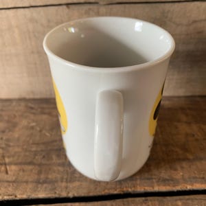 K&ouml;nnte beinhalten: Wei&szlig;er Keramikbecher mit gebogenem Henkel. Der Becher hat ein gelbes Design an der Seite. Der Becher steht auf einer Holzoberfl&auml;che.