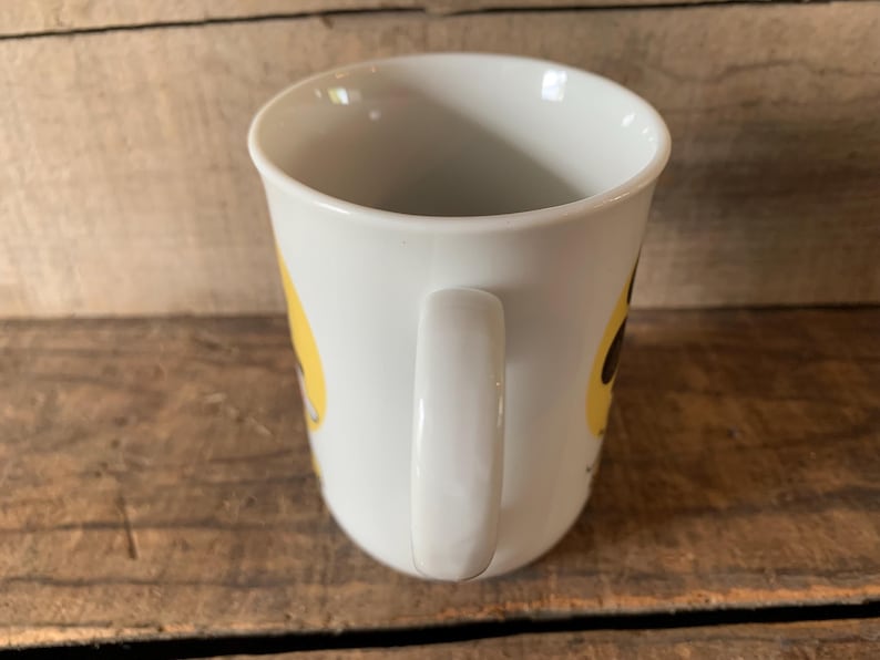K&ouml;nnte beinhalten: Wei&szlig;er Keramikbecher mit gebogenem Henkel. Der Becher hat ein gelbes Design an der Seite. Der Becher steht auf einer Holzoberfl&auml;che.