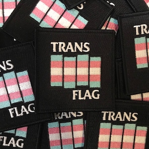 Peut inclure: Un ensemble de patchs en tissu noir avec le texte "TRANS FLAG" et un design stylisé du drapeau de la fierté transgenre.