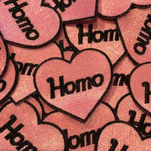 Könnte beinhalten: Ein Stapel rosa herzförmiger Patches mit dem Wort "Homo" in Schwarz auf jedem Patch.