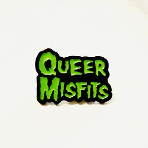 May include: Green and black enamel pin with the text "Queer Misfits" in a stylized font.