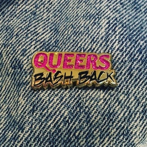 Könnte beinhalten: Eine goldfarbene Anstecknadel mit den Worten "QUEERS BASH BACK" in pinken Glitzerbuchstaben. Die Anstecknadel befindet sich auf einem blauen Jeansstoff.