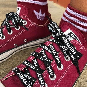 May include: Red Converse sneakers with black shoelaces that have the words "HE/THEY" printed on them. The sneakers are worn with red and white striped socks.