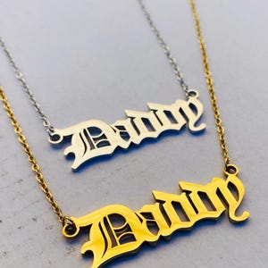 Puede incluir: Dos collares con el nombre "Danny" en fuente gótica. Uno es dorado y el otro plateado. Ambos tienen finas cadenas. Los collares se exhiben sobre una superficie gris claro.