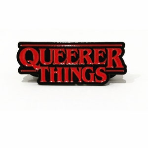 以下が含まれることがあります： 「ストレンジャー・シングス」の人気テレビ番組に似たスタイルで、「Queerer Things」というテキストが書かれた赤と黒のエナメルピンです。