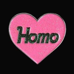 Può includere: Spilla a forma di cuore rosa con la parola "Homo" in lettere nere con glitter verde.