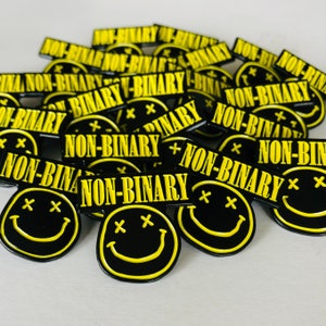 May include: A group of black and yellow enamel pins with a smiley face design and the text "NON-BINARY" on each pin.