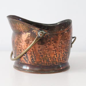 May include: A hammered copper coal scuttle with a wide, open top and a curved rim. The vessel features a metallic finish with visible oxidation and a brass swing handle attached to the sides. The base is circular and slightly flared at the bottom.
