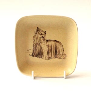 May include: A square, beige decorative plate featuring a detailed pencil drawing of a Yorkshire Terrier. The dog is depicted in a seated position with long, flowing fur. The plate is supported by a white stand.