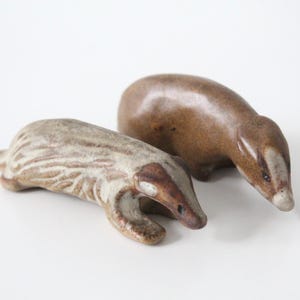 May include: Two small ceramic badger figurines. One figurine features a smooth, solid brown glaze. The second figurine has a textured, cream-coloured body with brown striped markings and a brown face, both sculpted in a low-profile, resting pose.