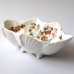 May include: A white ceramic bowl with a scalloped shell-like edge and three small feet. The interior features a colourful floral pattern with various small flowers, green leaves, and a prominent orange butterfly printed on the bottom surface.