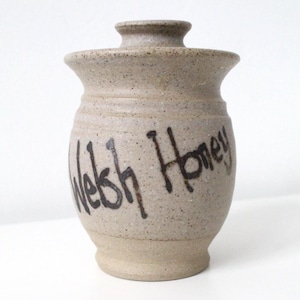 May include: A beige ceramic honey pot with a lid. The pot has a speckled texture and the words "Welsh Honey" in black script. The pot has a rounded body and a small, cylindrical lid.