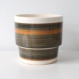 May include: A cylindrical ceramic planter featuring a horizontal striped pattern in shades of brown, grey, and orange. The vessel has a smooth, cream-coloured rim at the top and a matching cream-coloured base, with a stepped design along the body.