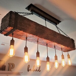 May include: A rustic wooden beam chandelier with eight Edison bulb lights hanging from black chains. The beam is dark brown with a rough, natural texture. The bulbs are lit and casting a warm glow.