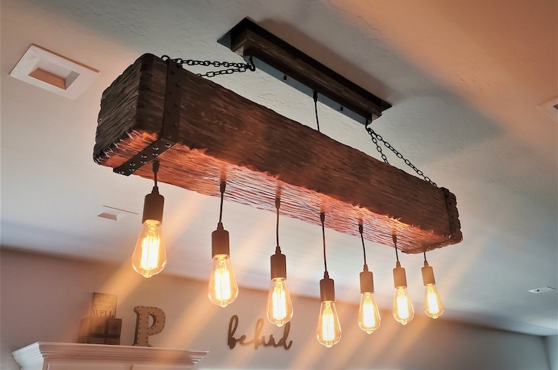 May include: A rustic wooden beam chandelier with eight Edison bulb lights hanging from black chains. The beam is dark brown with a rough, natural texture. The bulbs are lit and casting a warm glow.