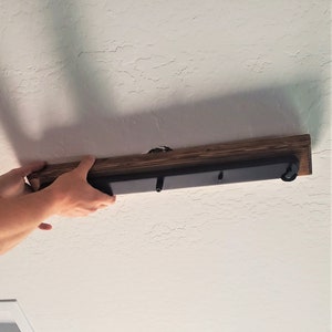 May include: A person installing a wooden ceiling light fixture with black metal accents. The fixture has three hooks for hanging light bulbs.