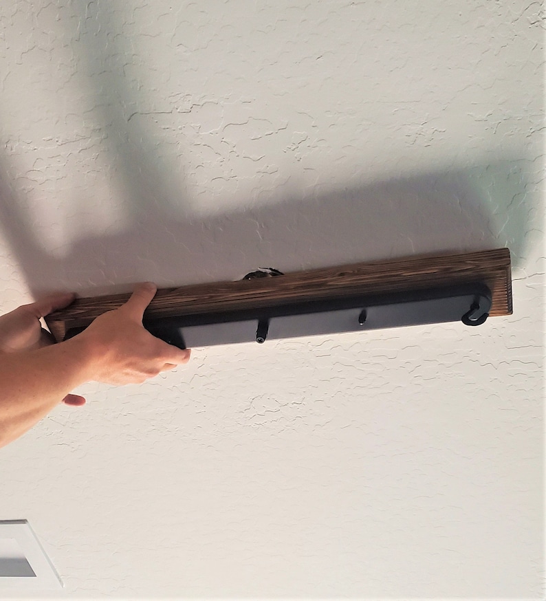 May include: A person installing a wooden ceiling light fixture with black metal accents. The fixture has three hooks for hanging light bulbs.
