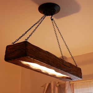 Può includere: Un lampadario da soffitto in legno rustico con forma rettangolare e una fonte di luce che corre al centro. Il lampadario è sospeso al soffitto da quattro catene nere.