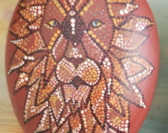 May include: A red, round stone painted with a lion's face using a dot technique. The lion's mane is depicted with black, white, and gold dots. The background is a red-brown color.