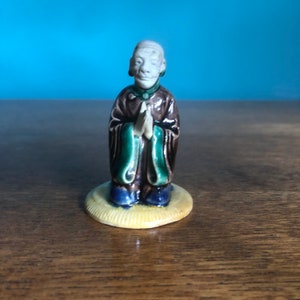 May include: A small ceramic figurine of an Asian man in a brown robe with green and blue detailing. The figure has hands clasped in prayer and stands on a yellow circular base. A collectible item.