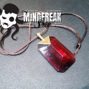 Może przedstawiać: Naszyjnik z czerwonym kryształowym wisiorkiem ze złotą górą i brązowym sznurkiem. Wisiorek w głębokim czerwonym kolorze ma fasetowany wzór. Naszyjnik to prawdopodobnie dodatek do cosplayu.