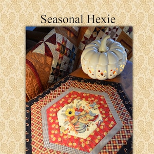 May include: A hexagonal table runner with a autumn-themed design featuring pumpkins, sunflowers, and leaves. The runner is made of patchwork fabric in shades of red, black, white, and yellow. The text "Sew'n Wild Oaks By Lynn Wilder" is visible in the bottom left corner of the image.