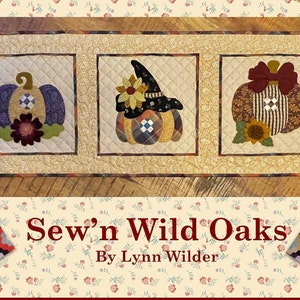 May include: A quilted table runner featuring three square panels with pumpkin designs. The pumpkins are in various colors and patterns, with floral and bow accents. The text "Sew'n Wild Oaks By Lynn Wilder" is displayed below the runner.