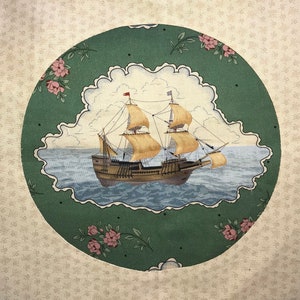 Op de afbeelding: Een groene en witte stoffen cirkel met een schip dat op een blauwe zee vaart. Het schip heeft bruine zeilen en een witte romp. De stoffen cirkel is omgeven door een witte achtergrond met een bloemenpatroon.