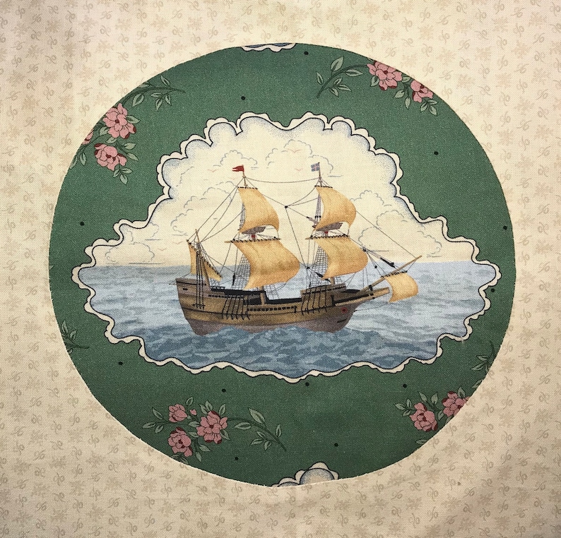 Op de afbeelding: Een groene en witte stoffen cirkel met een schip dat op een blauwe zee vaart. Het schip heeft bruine zeilen en een witte romp. De stoffen cirkel is omgeven door een witte achtergrond met een bloemenpatroon.