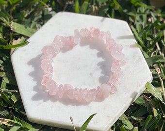 May include: A pink rose quartz chip bracelet on a white hexagonal surface. The bracelet is made of small, irregular pieces of rose quartz.
