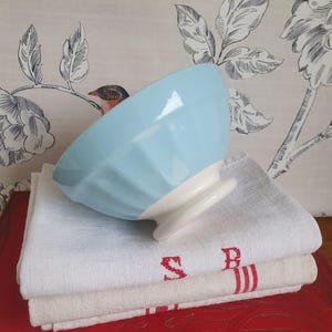 Może przedstawiać: Jasnoniebieska ceramiczna miska z żebrowaną powierzchnią zewnętrzną i białą podstawą, spoczywająca na stosie dwóch złożonych białych i beżowych lnianych serwetek z czerwonymi haftowanymi literami i paskami, umieszczona na czerwonej powierzchni.