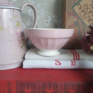 May include: A pink bowl with a white base sits atop a stack of vintage items, including a red book and a linen towel with red stripes and the letters "SB". A pink pitcher with floral designs is visible.