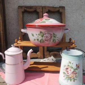 Pode incluir: Uma coleção de itens de cozinha vintage em esmalte. Inclui uma panela rosa e branca com detalhes de rosas, uma cafeteira rosa com acabamento dourado e um balde azul claro e branco com um design floral. Os itens estão dispostos em uma prateleira de madeira.