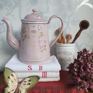 Könnte beinhalten: Eine altrosa Emaille-Teekanne mit floralen Details steht auf einem Stapel weißer Leinen mit roter Schrift. Ein Holzschmetterling und ein kleines Glas mit Holzutensilien sind ebenfalls vorhanden, was eine charmante, rustikale Ästhetik erzeugt.