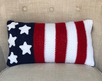 May include: A crocheted pillow in the shape of the American flag. The pillow is red, white, and blue with white stars on a blue field. The pillow is sitting on a beige armchair.