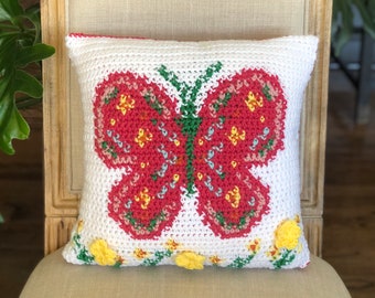 Op de afbeelding: Wit gehaakt kussen met een rood vlinderontwerp. De vlinder heeft gele en groene accenten. Gele bloemen en groene ranken versieren de onderrand. Het kussen ligt op een beige stoel.